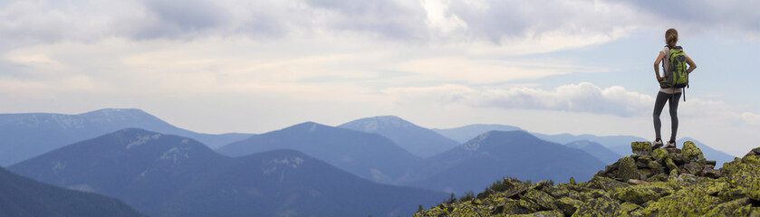 Back view of young slim girl with backpacks standing on rocky mountain top against bright blue...