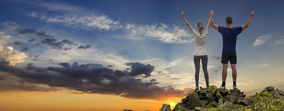 Panorama of young couple, athletic boy and slim girl standing with raised arms on rocky mountain top enjoying breathtaking summer mountain view. Tourism, traveling and healthy lifestyle concept.