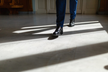 Obraz premium Legs of a man in an expensive suit and black shoes during a step. A person walks along the corridor of the living room or hotel marble floor under the sunlight. Legs close up. Business concept