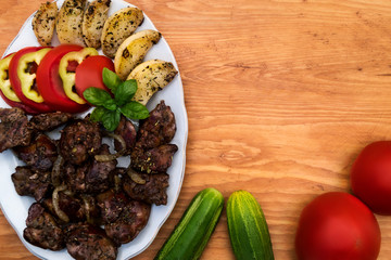 Fried chicken liver with apples and onions on a plate, decorated with slices of tomato and sweet pepper, next to the table cucumbers and tomatoes, wooden background.