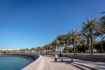 Doha, Qatar - Jan 9th 2018 - Tourists walking close to the bay with palm tree shades in a blue sky...