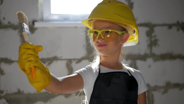 childhood, construction, architecture, building and people concept - smiling little girl in protective yellow helmet and safety glasses with brush for paint