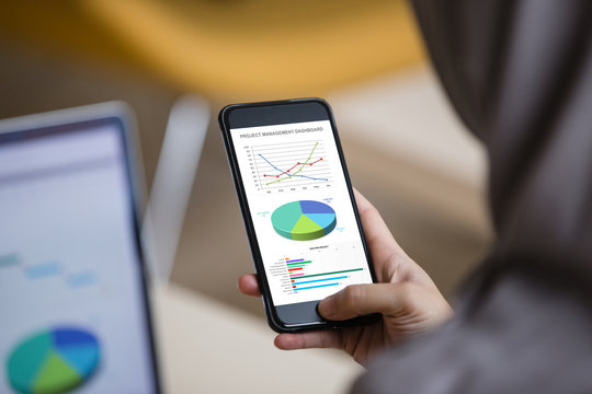 Project management dashboard concept. Hijab woman holding phone with excel project dashboard on screen with laptop background.