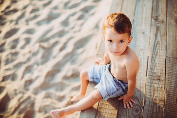 Fototapeta na wymiar The theme is a child and summer beach vacation. A small Caucasian boy sits sideways on a wooden pier and looks at the camera on a sandy beach and a pond, a river. With bare legs in blue denim shorts