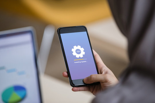 Hijab woman holding smartphone doing installing update process with gearbox percentage progress and loading bar with laptop background