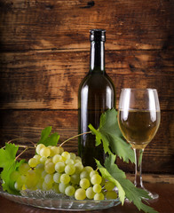 Glass with white wine, black bottle and grapes in bowl decorated with vine. Retro and vintage wooden background. Food and drink decoration concept. Close up, selective focus