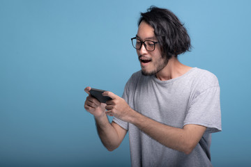 Excited Young bearded man playing mobile games in blue background.