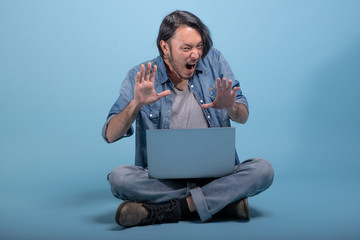 Young bearded man sit down on floor using computer in blue background.