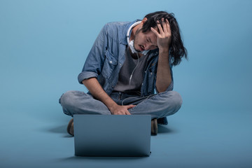 Young bearded man sit down on floor using computer in blue background.