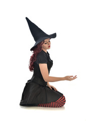 Fototapeta premium full length portrait of red haired girl wearing black witch costume and pointy hat. seated pose, isolated on white studio background.