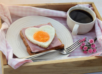 Heart shape sunny side up egg with bread,ham and coffee on pink check fabric background in wood tray, breakfast for valentine or mothers day