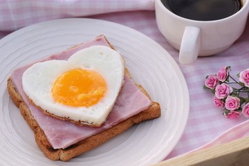 Fototapeta premium Heart shape sunny side up egg with bread,ham and coffee on pink check fabric background in wood tray, breakfast for valentine or mothers day