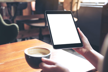 Mockup image of woman's hands holding black tablet pc with blank white screen with coffee cup on wooden table in cafe
