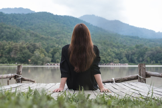 Closeup image of an asian woman sitting alone on an old wooden pier by the river with sky and mountain background