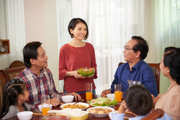 Pretty young Asian woman bringing bowl of salad to dinner table