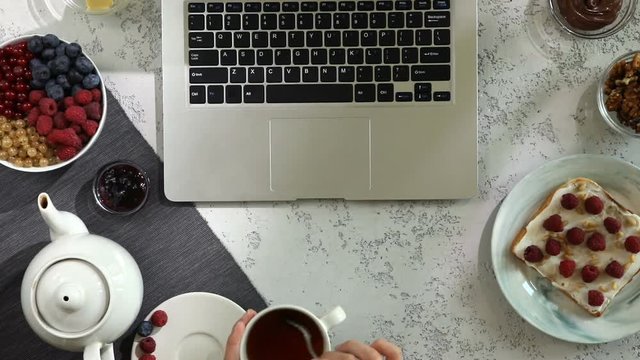 Top view of woman hands on workspace with coffee break with laptop, bread toasts with berries and cup of black tea composition with shadows and wind. Flat lay, view from above