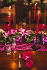 Wooden wedding table decorated with red candles, pink cloth and purple orchids. Romantic family dinner in evening