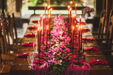 Wooden wedding table decorated with red candles, pink cloth and purple orchids. Romantic family dinner in evening