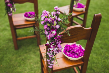 Brown wooden chairs standing on grass on wedding ceremony decorated with pink and purple flowers. Plate with flower petals