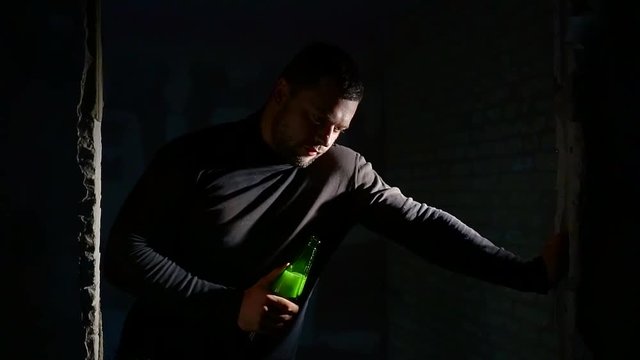 drunk homeless man is holding bottle and leaning on door hole in dark ruined building