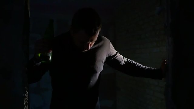 drunken man is staggering inside a door hole in a dark basement, holding bottle in hand