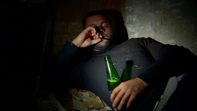 drunken unshaven dirty bum man is lying in a darkness with cigarette and bottle with alcohol beverage