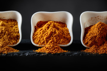 Small white ceramic bowls of Indian spices on dark background .