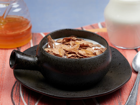 A bowl of oat bran flakes with milk and honey set on a red napkin, on a blue tablecloth, with a jar of honet and a jug of milk