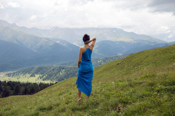 Naklejka na ściany i meble woman in a blue long dress stands on top of a mountain in the summer on a green meadow
