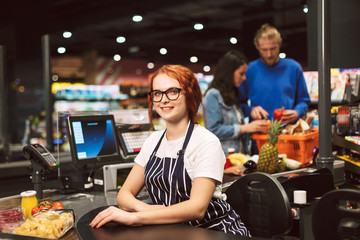 Young smiling cashier in eyeglasses and striped apron happily looking in camera while working in...