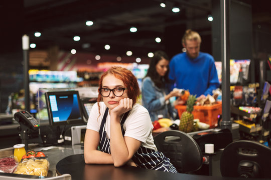 Young boring cashier in eyeglasses and striped apron thoughtfully looking in camera while working in modern supermarket with customers on background