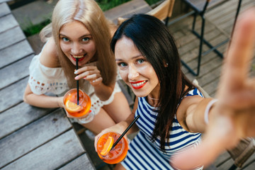 Female friends taking selpfie photos loking at camera and enjoying fresh drinks. Top view of girls toasting each other with aperol spritz cocktails at the wooden table.