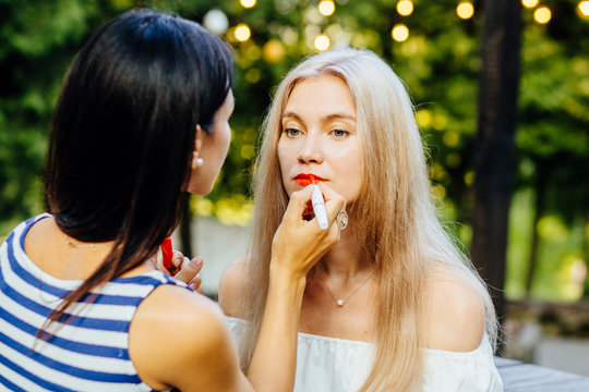 Young brunette woman painting lips for her girl friend with red lipstick outdoor being on the street. Summer holidays, people, friendship and party time concept.