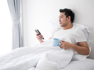 Happy Asian man using smartphone and drinking coffee on bed, man lifestyle on weekend concept.