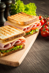 Close-up of two sandwiches with bacon, salami, prosciutto and fresh vegetables on rustic wooden cutting board. Club sandwich concept.