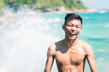 Happy young Thai man contagiously laughs on a beach having behind sunny seascape and wave splash. A...