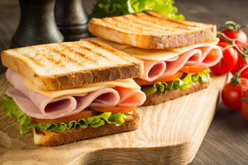 Close-up of two sandwiches with bacon, salami, prosciutto and fresh vegetables on rustic wooden cutting board. Club sandwich concept.