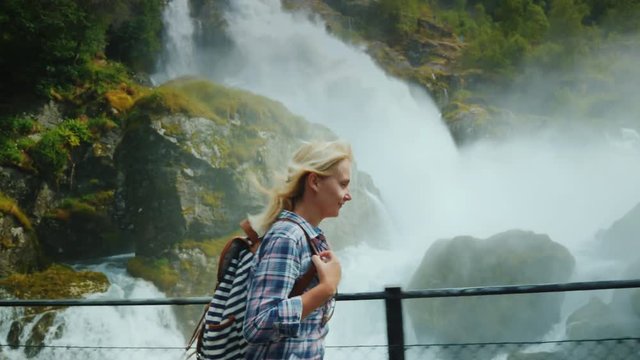An active woman with a backpack walks across the bridge against a waterfall on the way to the Briksdal Glacier. Drops of water fly in her face. Traveling in Norway