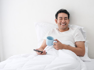 Smiling Asian man using smartphone and drinking coffee on bed, man lifestyle on weekend concept.