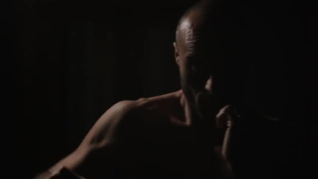 A lone man is boxing in a dark room, watching the camera, with an hard light moving around
