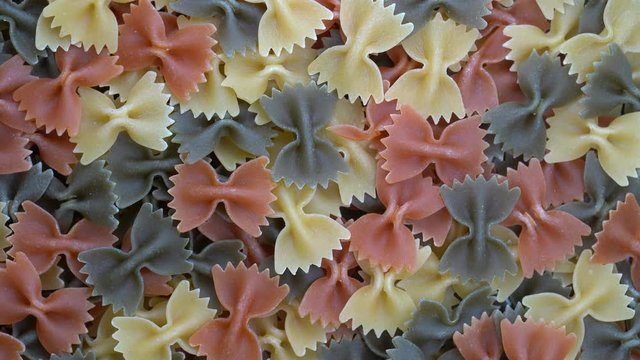 Multicolored pasta background, rotating, top view. Rotation of colored farfalle pasta. Heap of uncooked pasta. Italian food close up. Delicious italian farfalle tricolor pasta, food background