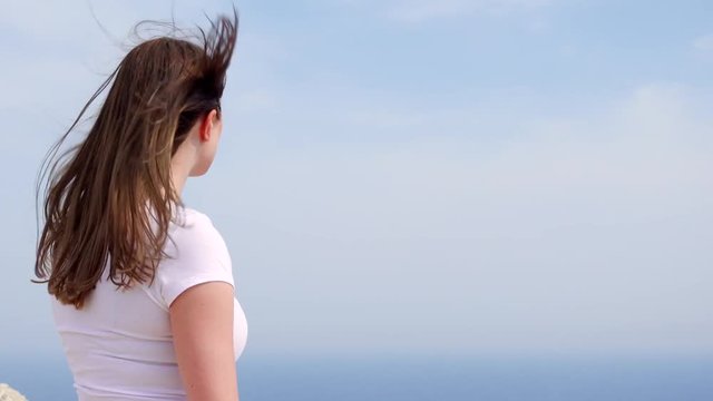 Smiling young woman in white t-shirt standing at edge of cliff on sunny day. Carefree female looking at breathtaking view of blue Mediterranean sea. Concept of freedom and inspiration