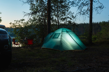 Night landscape with a tent in the forest near lake. The light from the lantern in a tent. Car and portable table and chairs, green tourist tent. Romantic evening with a tent at night.