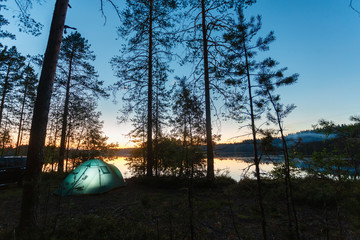 Night landscape with a tent in the forest near lake. The light from the lantern in a tent. Car and portable table and chairs, green tourist tent. Romantic evening with a tent at night.