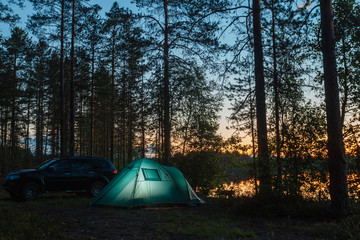 Night landscape with a tent in the forest near lake. The light from the lantern in a tent. Car and portable table and chairs, green tourist tent. Romantic evening with a tent at night.