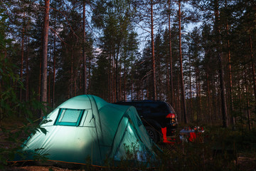 Night landscape with a tent in the forest near lake. The light from the lantern in a tent. Car and portable table and chairs, green tourist tent. Romantic evening with a tent at night.