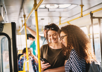Two best young sweet girl friends standing in a bus and looking in a telephone.