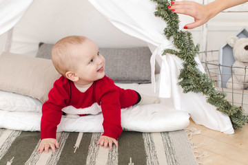 Lovely baby in red clothing smiling and looking away while lying in small tent decorated with conifer branches