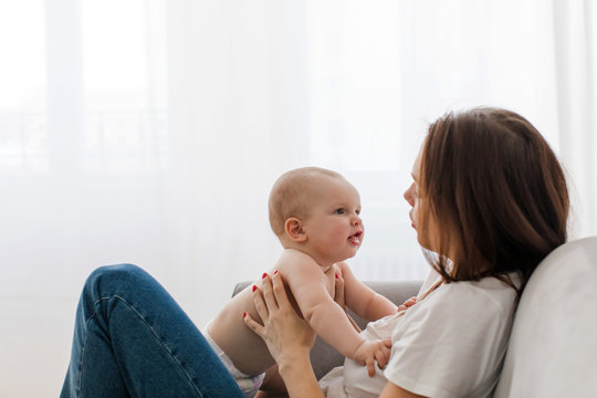 Side View Of Young Mother Sitting On Sofa With Baby On Knees And Talking On Background Of White Wall