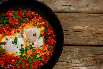 Homemade breakfast shakshuka - fried eggs, onion, bell pepper, tomatoes and parsley in cast-iron frying pan on rustic wooden table. Jewish cuisine. Top view